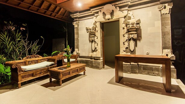 A traditional wooden bench and sofa set with a vase of flowers, a wooden table, and a stone wall with carved details. - Outpost Ubud Coliving Suites (Ubud)