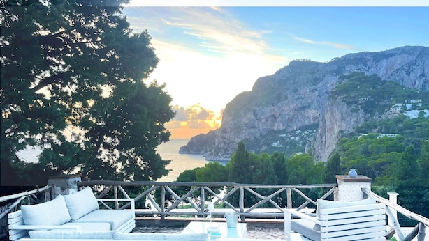 Une terrasse dotée de mobilier blanc, d’un sol en pierre et offrant une vue sur un paysage côtier avec des falaises et une étendue d’eau.