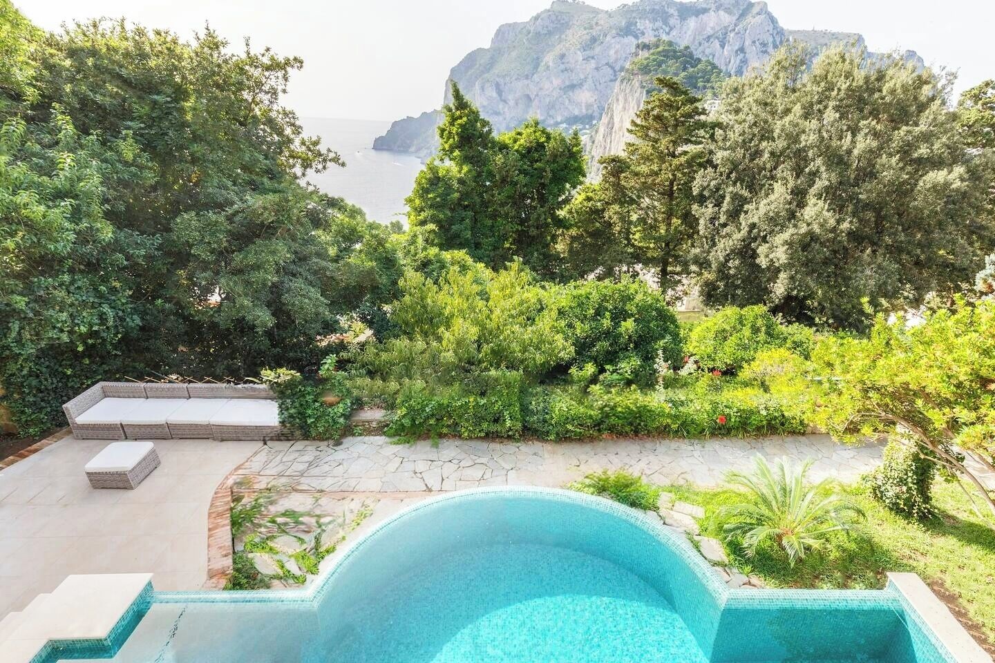 A swimming pool with a stone deck, surrounded by lush greenery and a view of the sea and mountains.
