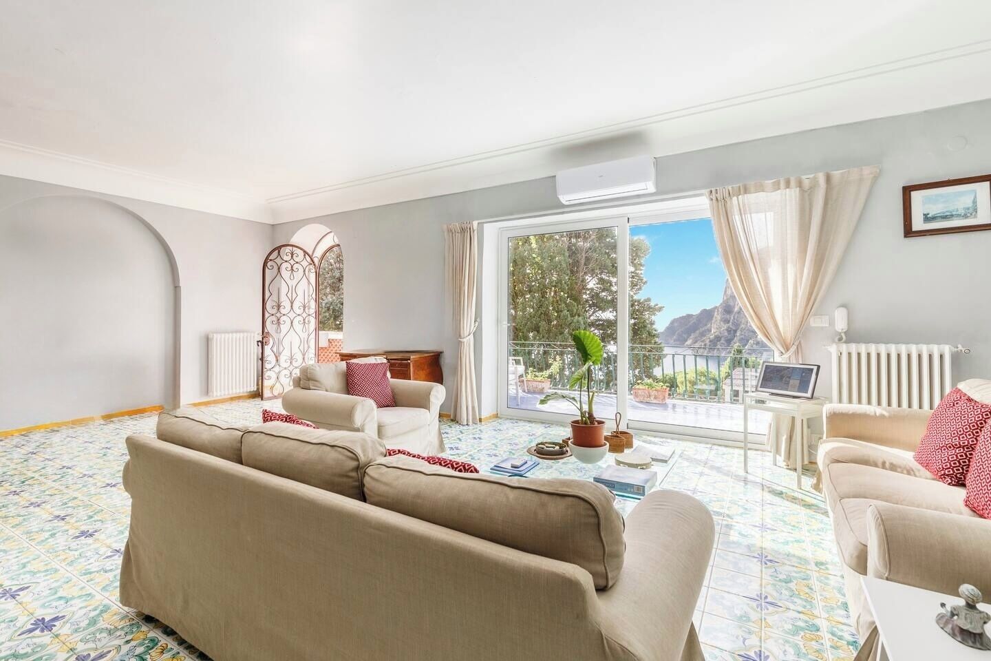 Un salon spacieux avec un grand canapé, un canapé plus petit et une table basse. Il y a un balcon avec vue sur les montagnes et une plante en pot sur le rebord de la fenêtre.