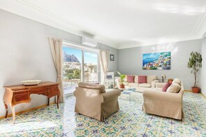 A spacious living room with a balcony, a wooden desk, two beige armchairs, a sofa, and a colorful tiled floor.