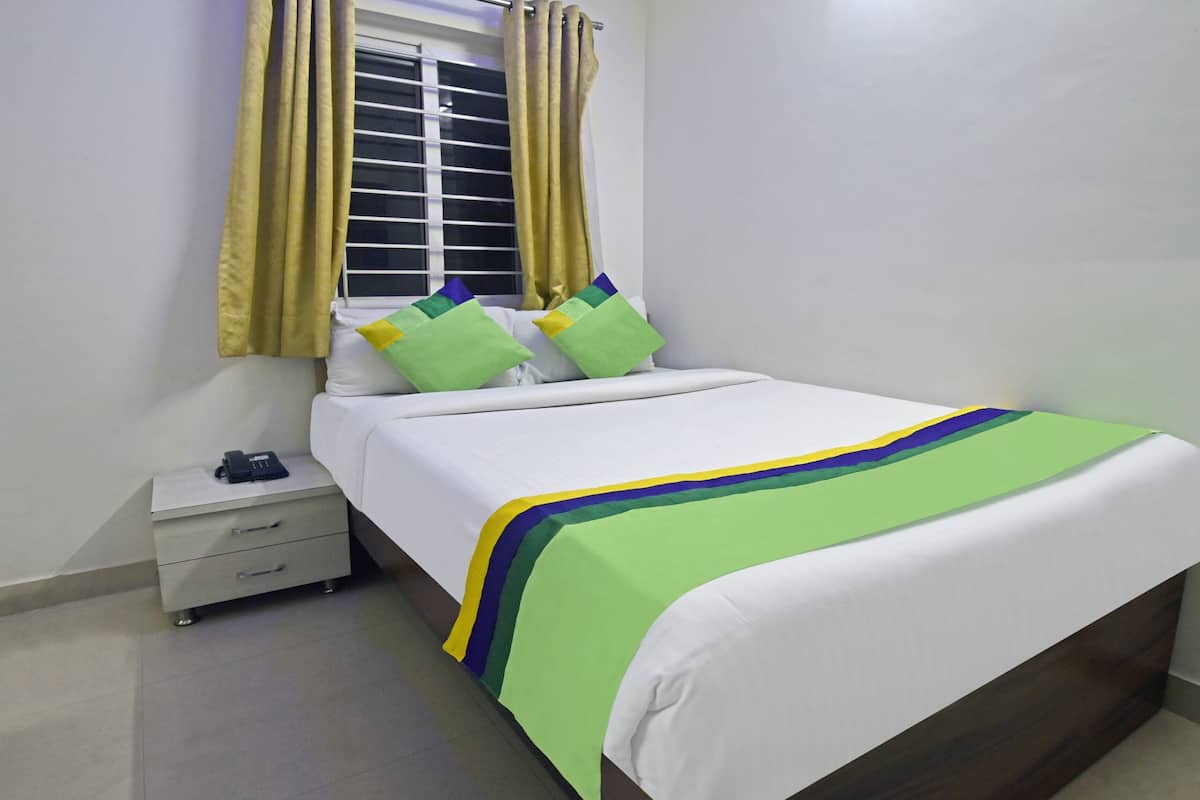 A neatly made bed with a green and blue striped bedspread, a white pillow, and a green pillow. There is a bedside table with a phone and a lamp. The room has white walls and a window with yellow curtains.