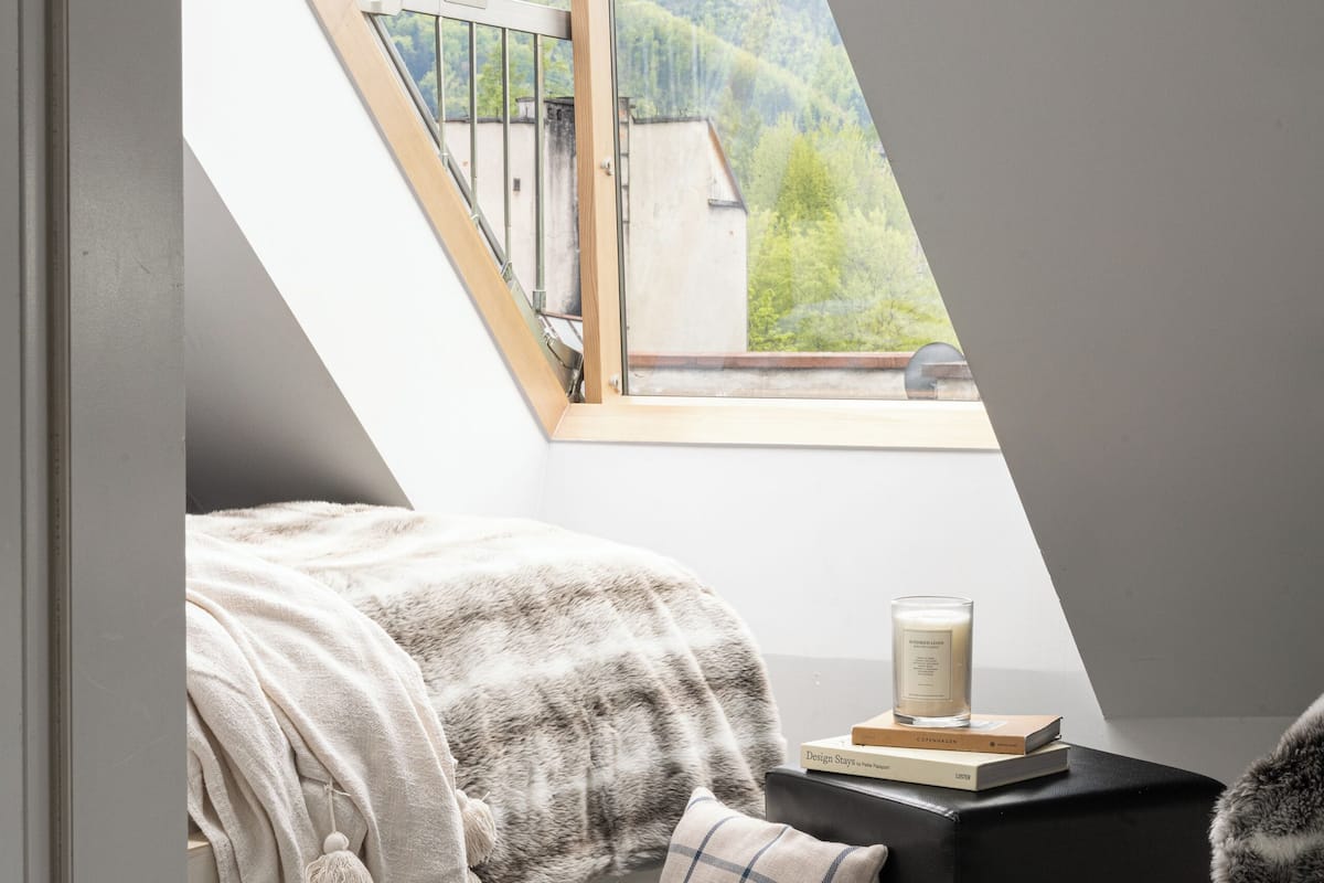 A bedroom with a skylight, a bed with a blanket, a pillow, a bedside table with a book and a candle, and a view of mountains.