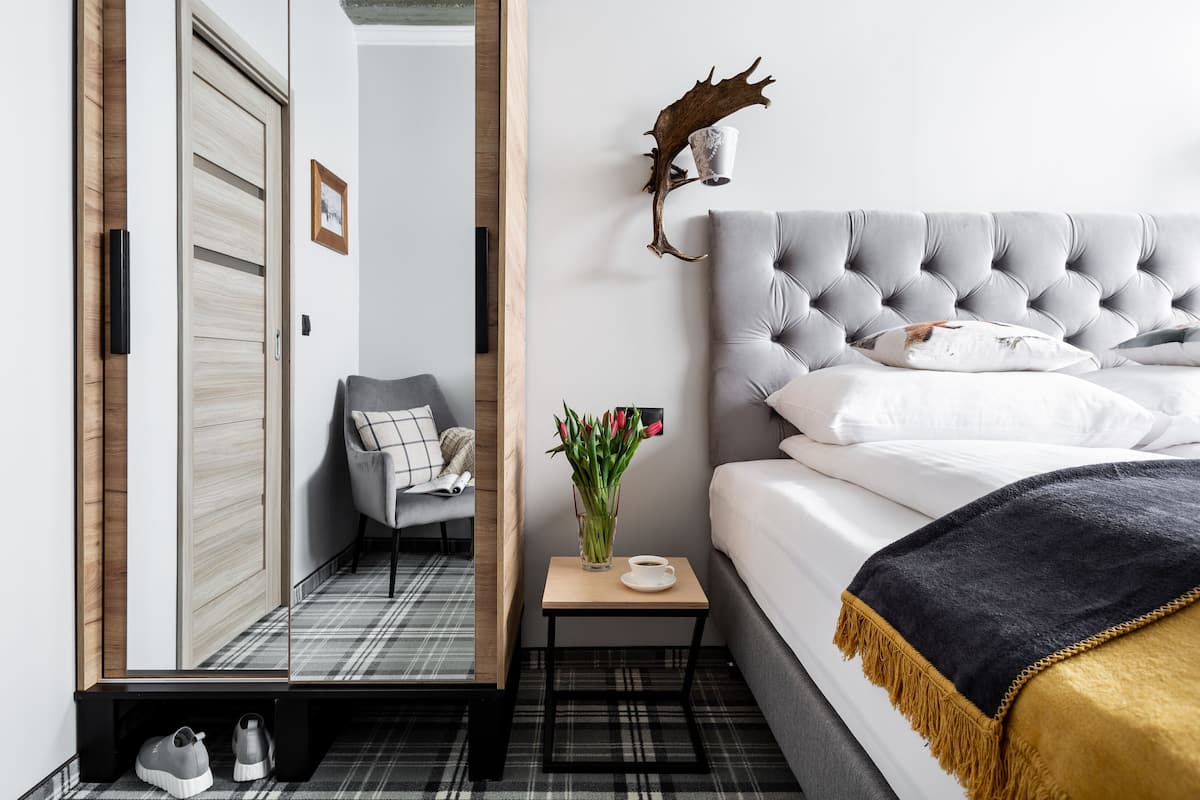 A modern hotel room with a grey tufted headboard, a small bedside table with a cup and flowers, a grey armchair, and a mirror reflecting the room.
