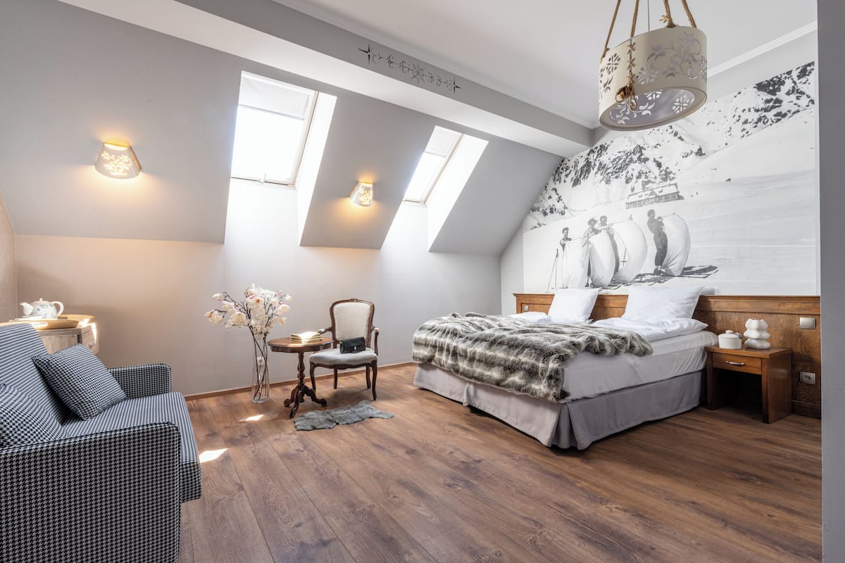 A bedroom with a bed, a chair, a small table, and a wall-mounted light fixture.