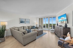 A living room with a sectional sofa, a flat-screen TV, and a view of the ocean through large windows.