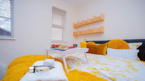 A neatly made bed with a yellow and white floral bedspread, a bedside table with a glass of orange juice, a bowl of fruit, and a bottle of lotion.