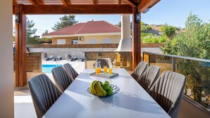A covered outdoor dining area with a pool and a view of a building with a red-tiled roof. - Panthea Villa in Ialysos (Ιαλυσός)