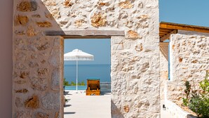 A stone archway leading to a beach area with a wooden chair and a white umbrella. - Terra Lucis Mountain Residences/ Mare Villa (Zakynthos)