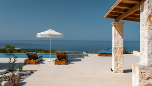 Zona de piscina amb gandules i un parasol blanc, amb vista al mar blau i transparent.