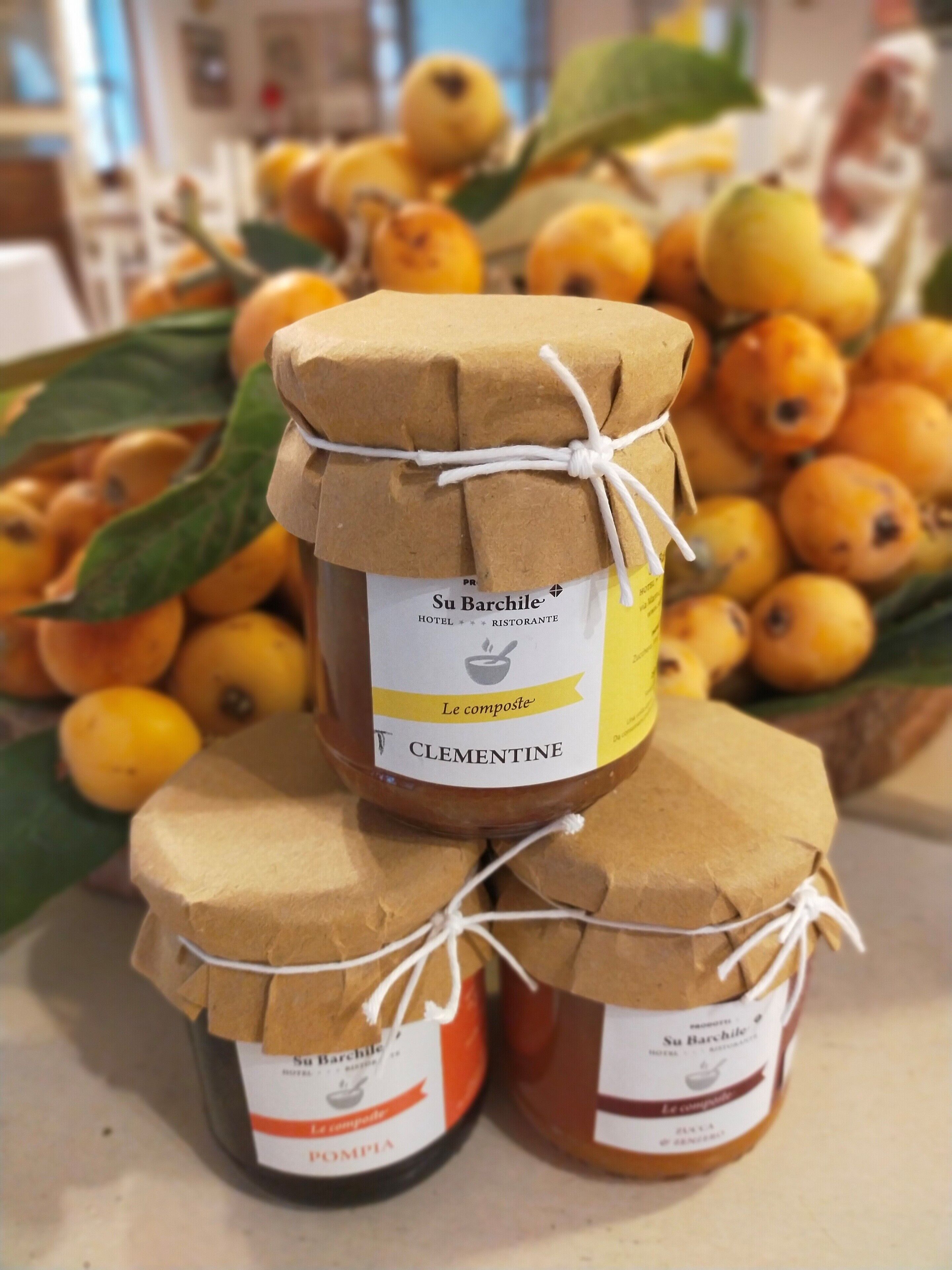 Tres frascos de mermelada de Clementine con etiquetas de papel marrón atadas con cuerda blanca, sobre una mesa con más frascos y limones al fondo.