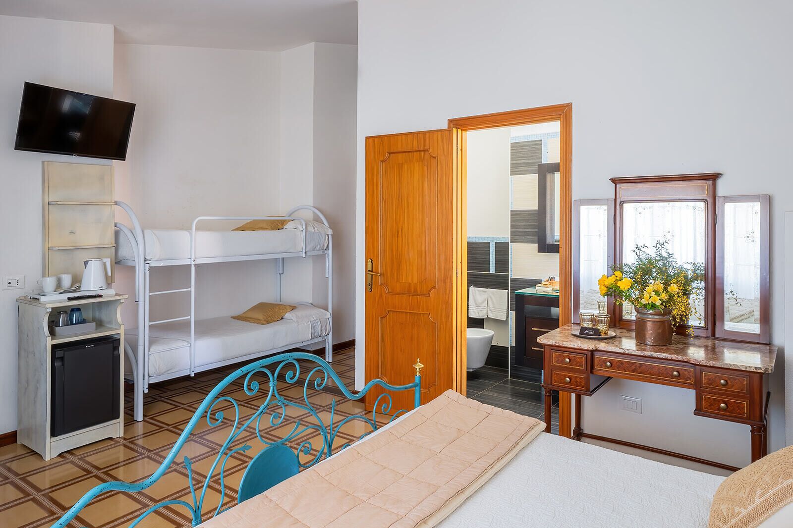 Quarto com beliches, pequeno espaço de cozinha, cómoda de madeira e televisão.