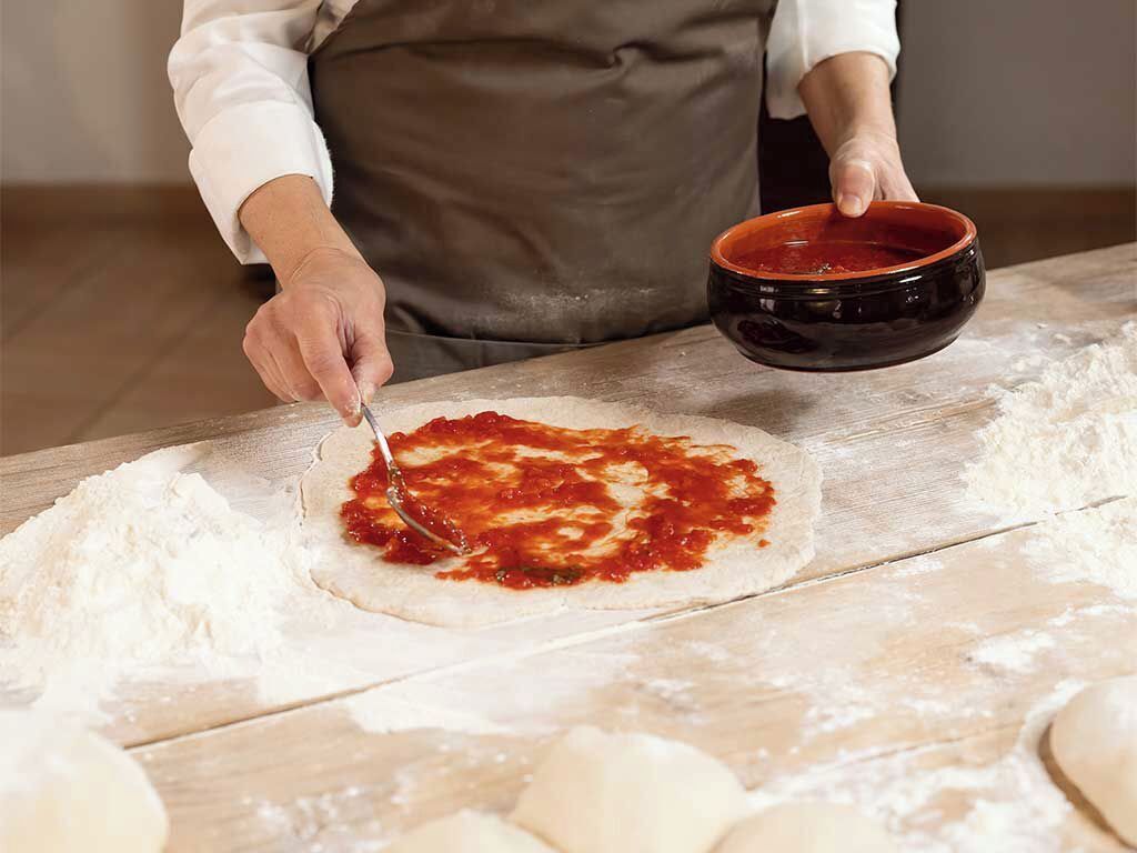 En person laver pizzadej på en træoverflade med mel og en skål tomatsovs.