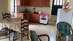 Een kleine keuken met houten kastjes, een eettafel met stoelen, een koelkast en een raam met gordijnen.