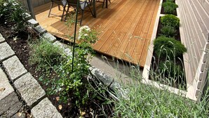 Een overdekt terras met een houten terras, een eettafel met stoelen, en een kleine tuin met planten en een stenen rand.