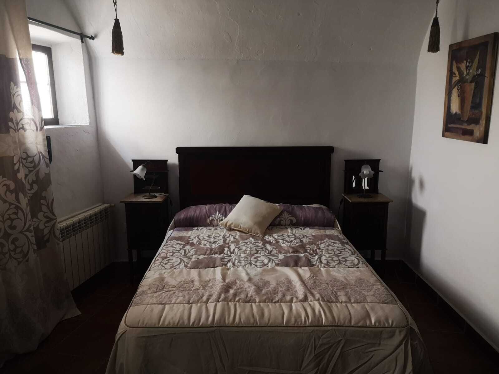 Un dormitorio con una cama grande, dos mesitas de noche con lámparas, un radiador y una ventana con cortinas.