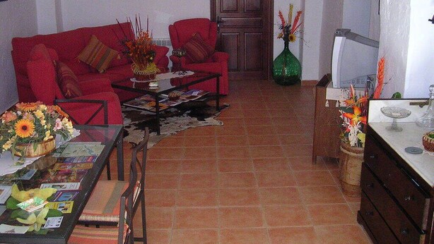 A cozy living room with a red sofa, a coffee table, and a dining table with flowers. There is a door and a green vase with flowers on the right side. - Los Gaitanes Rural House (Valverde de Leganés)