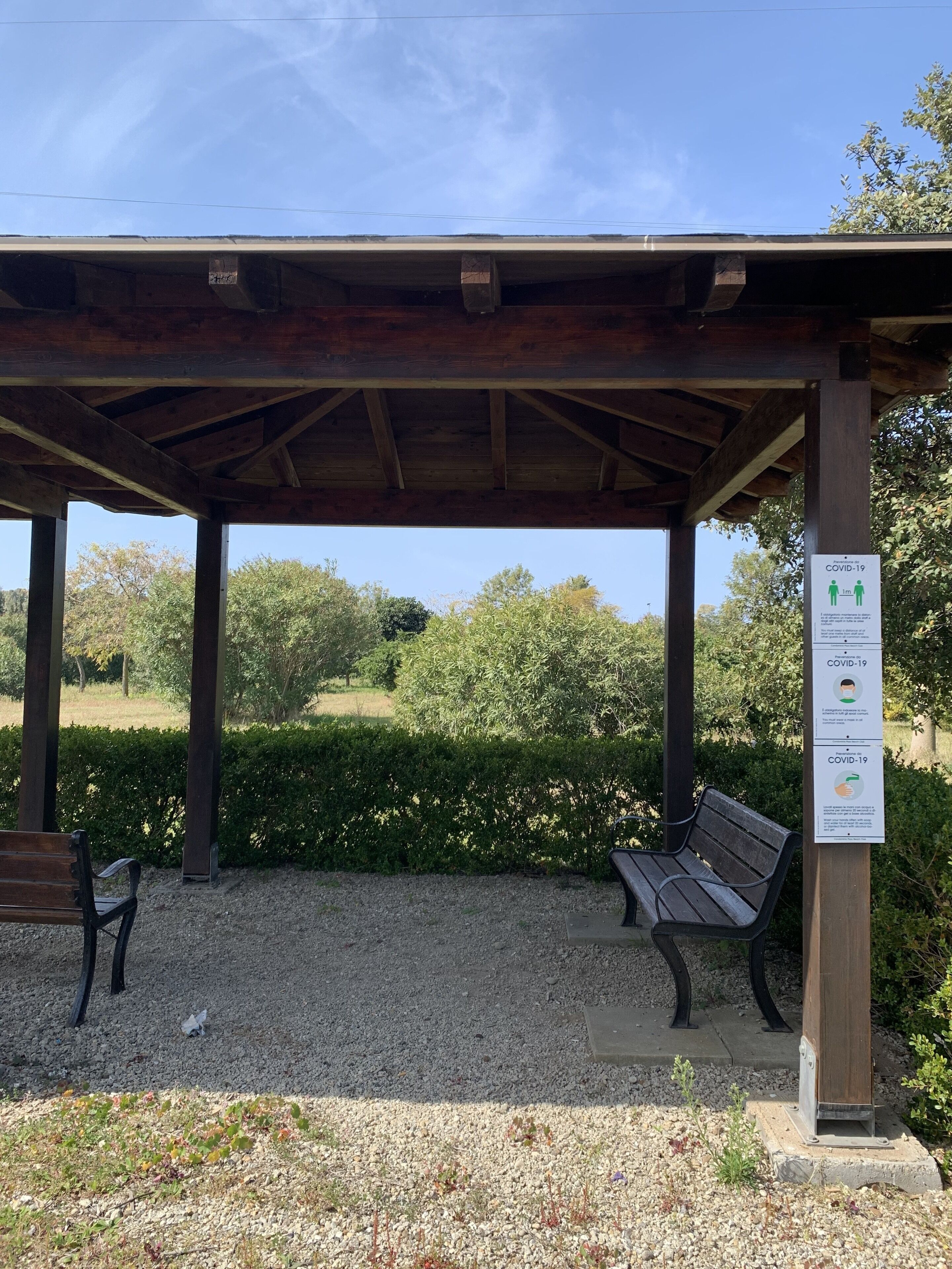 A covered outdoor seating area with benches, a gravel ground, and a sign with COVID-19 guidelines.