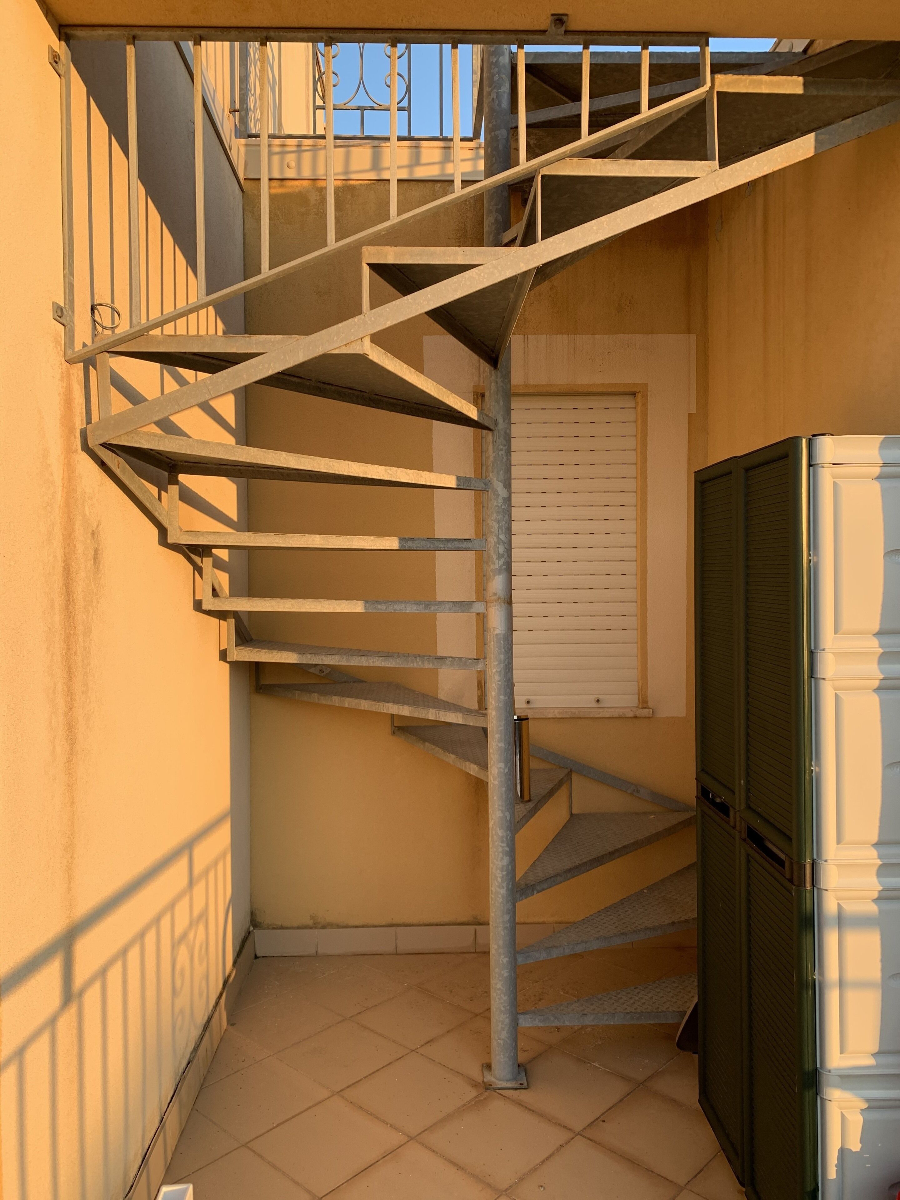 A metal spiral staircase in a narrow corridor with a tiled floor, a closed shutter door, and a refrigerator.
