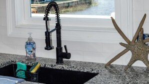 Eine Küche mit Granitarbeitsplatte, einem schwarzen Wasserhahn, einer Meerstern-Dekoration und einem Fenster mit Blick auf einen Steg und Wasser.
