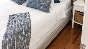 Ein ordentlich bezogenes Bett mit grauem Kopfteil, weißen und blauen Kissen sowie einer grauen Tagesdecke. Darüber an der Wand befinden sich drei gerahmte Bilder.