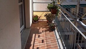 Un balcone con pavimento piastrellato, piante in vaso e ringhiera bianca.