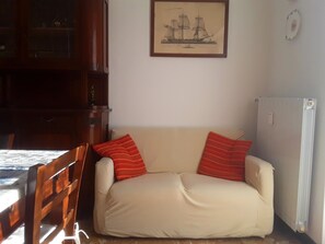 Ein Wohnzimmer mit einem beigen Sofa, zwei roten Kissen mit Streifenmuster, einem Holzstuhl, einem Bilderrahmen mit einem Schiffsbild an der Wand und einer Heizung.