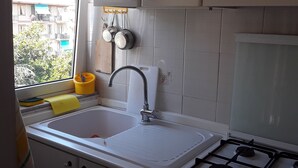 Una cucina con mobili bianchi, un lavello, un fornello e una finestra con vista su edifici e alberi.