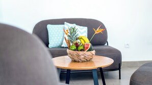 A room with a round wooden table, a woven fruit basket, and a grey sofa with a light blue pillow.