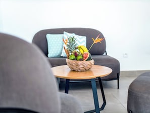 A room with a round wooden table, a woven fruit basket, and a grey sofa with a light blue pillow.