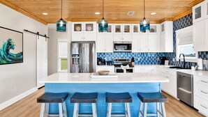 A modern kitchen with a large island, bar stools, stainless steel appliances, and a blue backsplash. - Luxury Coastal Retreat | Gulf Views | Private Elevator (Gulf Shores)