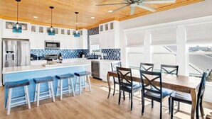 A modern kitchen with a wooden ceiling, a central island with bar stools, and a dining area with chairs. - Luxury Coastal Retreat | Gulf Views | Private Elevator (Gulf Shores)