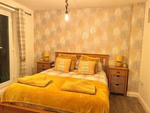 A bedroom with a bed, two bedside tables, a lamp on each table, a window with curtains, and a wall with a leaf pattern. - Sandy Bottom Apartment is a 1st floor open plan Appt on The Bay, Filey (Filey)