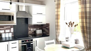 A modern kitchen with white cabinetry, a built-in oven, a microwave, and a gas stove. There is a dining area with a round table and white chairs. - Sandy Bottom Apartment is a 1st floor open plan Appt on The Bay, Filey (Filey)
