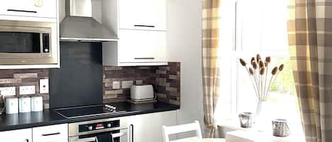 Uma cozinha moderna com armários brancos, forno embutido, micro-ondas e fogão a gás. Há uma área de jantar com uma mesa redonda e cadeiras brancas.