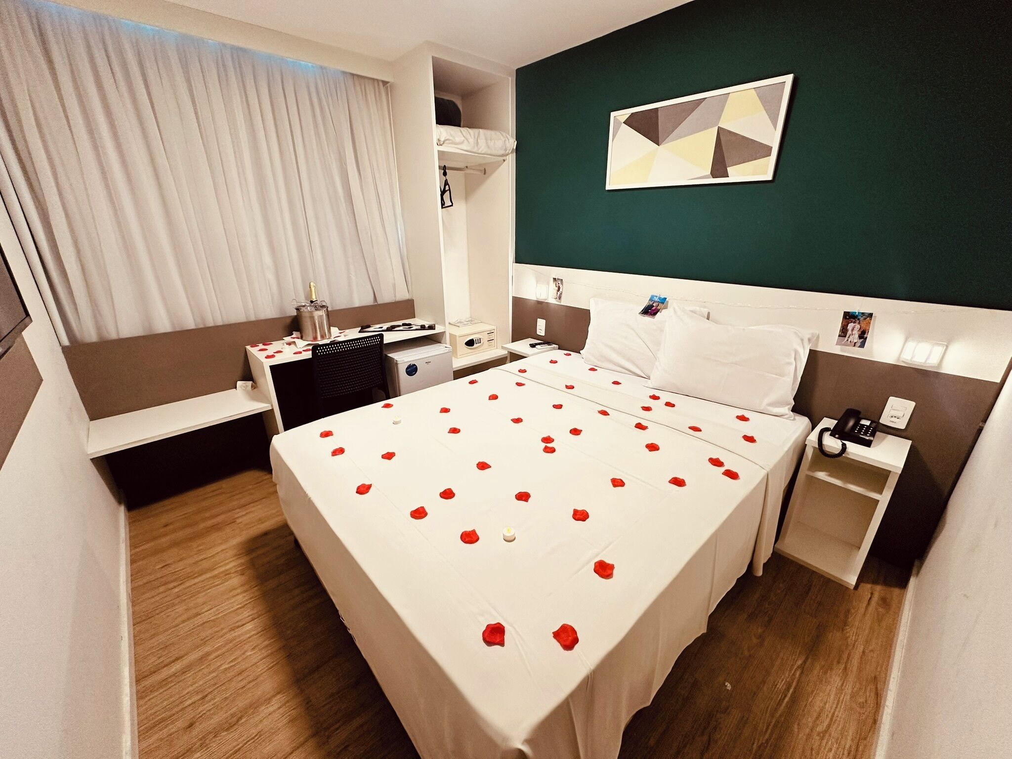 Una habitación de hotel con cama, mesitas de noche, escritorio, silla, ventana con cortinas y un cuadro en la pared.