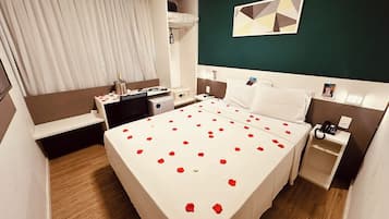Una habitación de hotel con cama, mesitas de noche, escritorio, silla, ventana con cortinas y un cuadro en la pared.