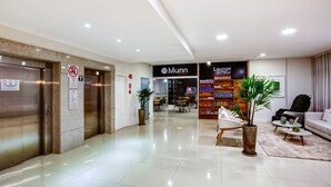Un vestíbulo de hotel con bar lounge, ascensores y una zona de estar con una mesita de café y plantas.