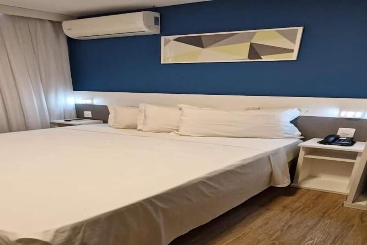 A hotel room with a large bed, a bedside table with a phone, and a wall-mounted air conditioner.