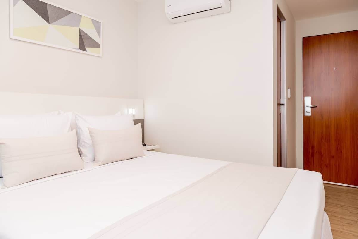 A hotel room with a bed, a bedside table, an air conditioning unit, and a wooden door.