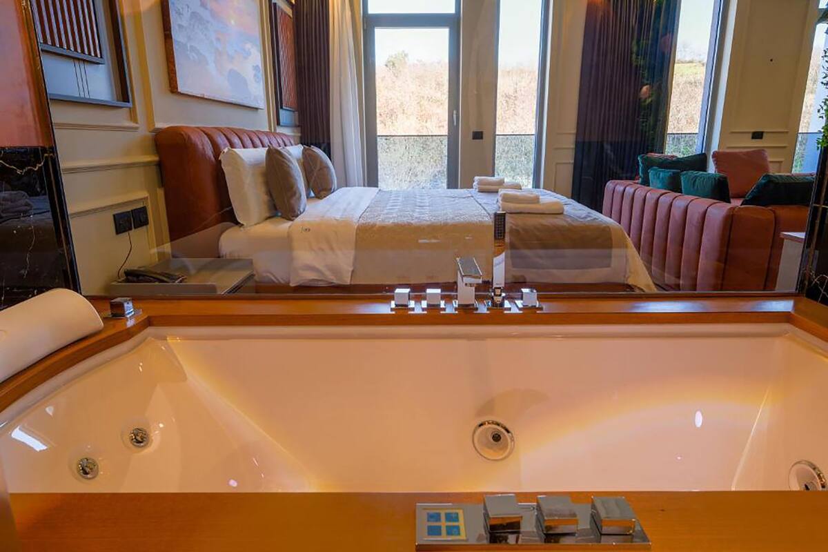 A hotel room with a large bathtub, a bed with a white and brown color scheme, and a view of the outdoors through large windows.