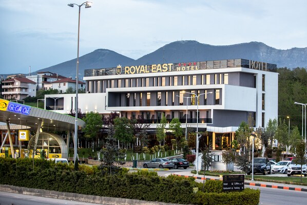 The Royal East Hotel with a modern design, a signboard indicating hotel, coffee, and parking facilities, and a view of the surrounding area with mountains in the background. - Royal East Resort (Farkë)