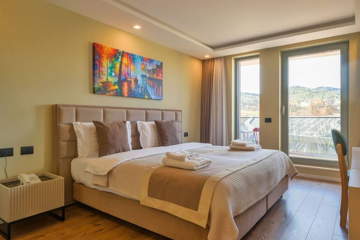 A modern hotel room with a large bed, a painting on the wall, a view of the outdoors, and a small table with a chair.