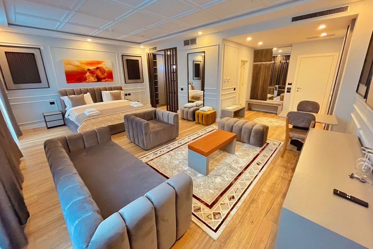 A modern hotel room with a large bed, a sofa, a coffee table, and a desk with chairs.
