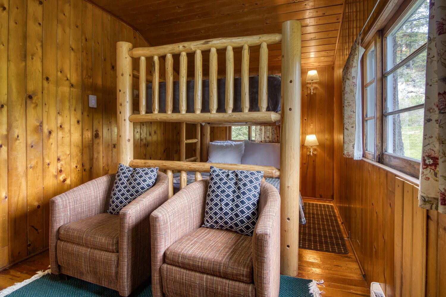 A cozy cabin interior with a wooden bed, two plaid armchairs, and a window with curtains.