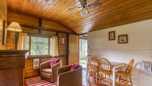 Interior acogedor de una cabaña con paredes de madera, un comedor con sillas de madera, un ventilador de techo y una ventana con cortinas.