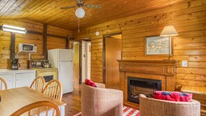 Interior acogedor de una cabaña con una mesa de comedor, sillas, chimenea, ventilador de techo y paredes de madera.