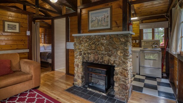 Un acogedor interior de cabaña con chimenea de piedra, vigas de madera, piso a cuadros y una kitchenette.