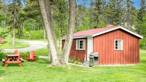 Una pequeña cabaña roja con techo rojo, una mesa de picnic y dos sillas Adirondack rojas al frente.