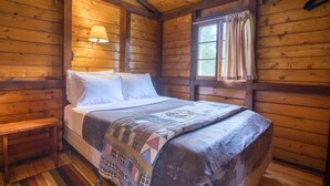 Un dormitorio de cabaña de madera con una cama, un banco, una ventana con cortinas y una lámpara.